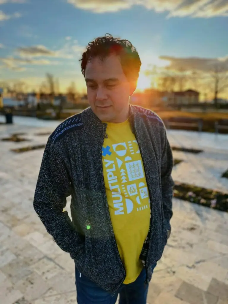 Man in a gray zip-up cardigan and yellow Multiply T-shirt standing on a stone plaza at sunset