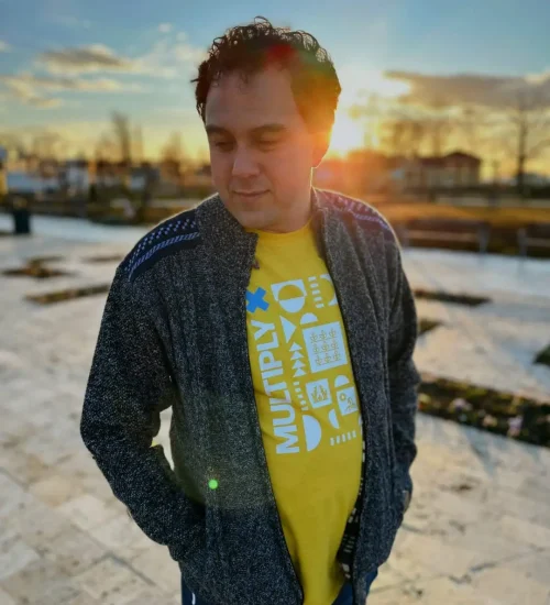 Man in a gray zip-up cardigan and yellow Multiply T-shirt standing on a stone plaza at sunset